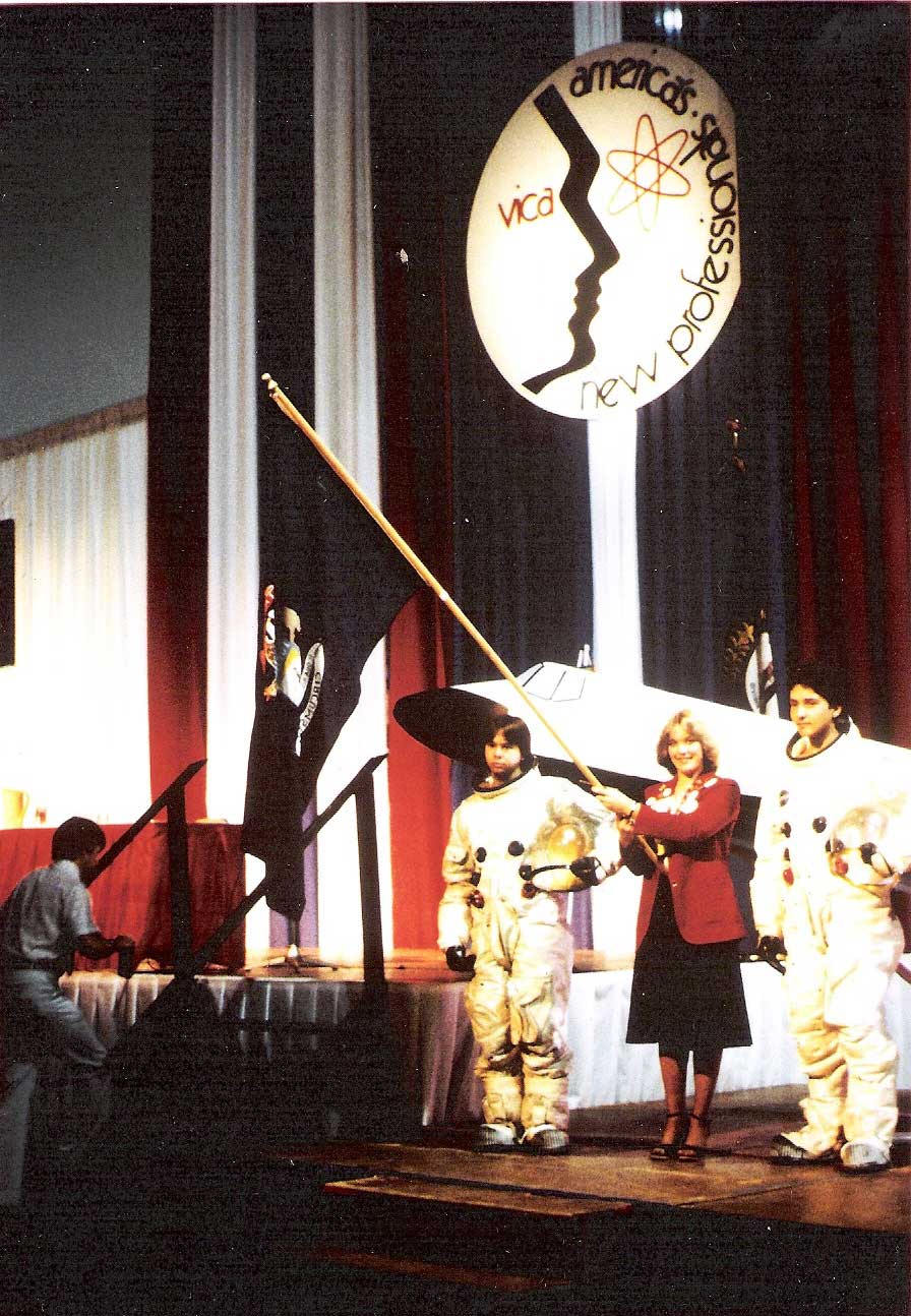 VICA students on stage with a flag and the sign saying America's New Professionals. Two men in space suits nearby}