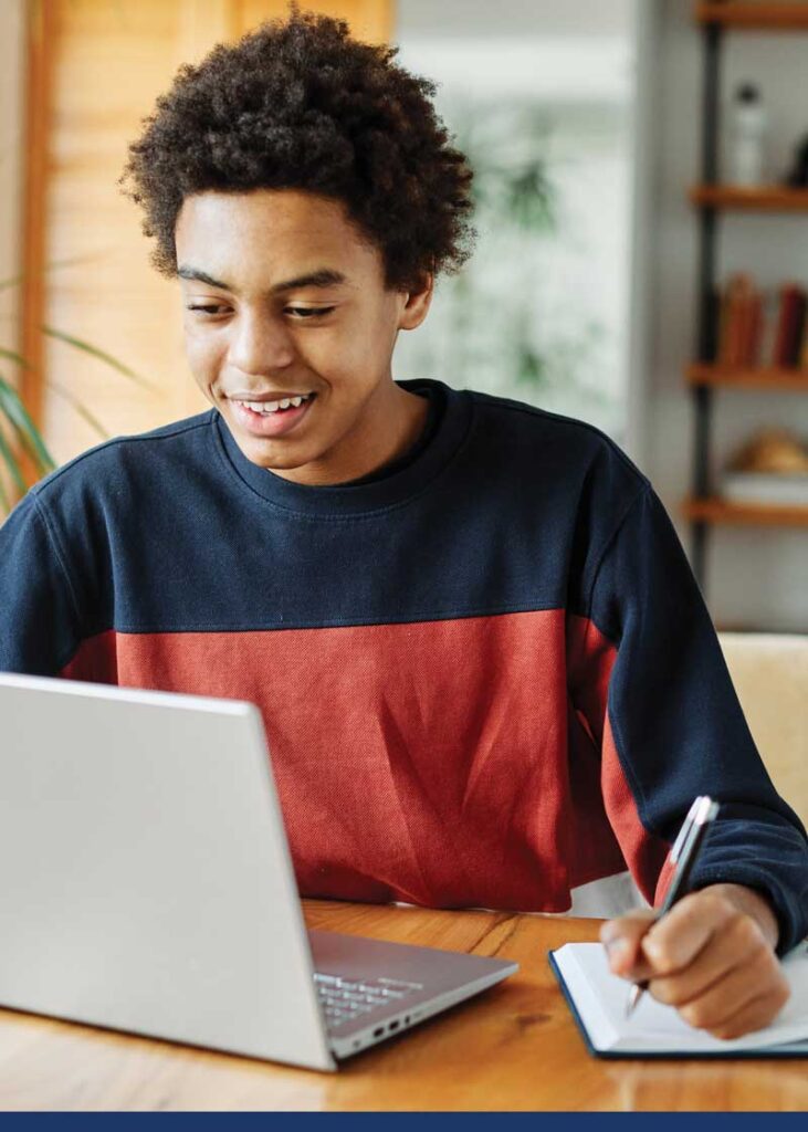 Young man smiling while looking at a laptop and writing in a notebook.