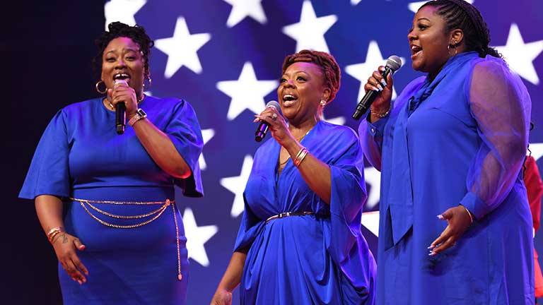 Three women in blue dresses in front of a U.S. flag image, singing