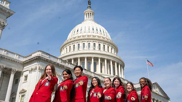 8 students wearing SkillsUSA red blazers stand in front of the U.S. Capitol pointing at the viewer.