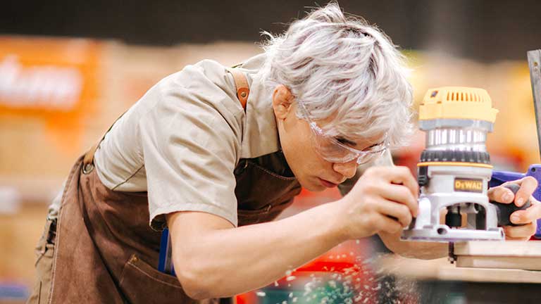 woodworker in safety glasses and a brown leather apron leans in with focused concentration, guiding a handheld router along the edge of a board as wood shavings fly.