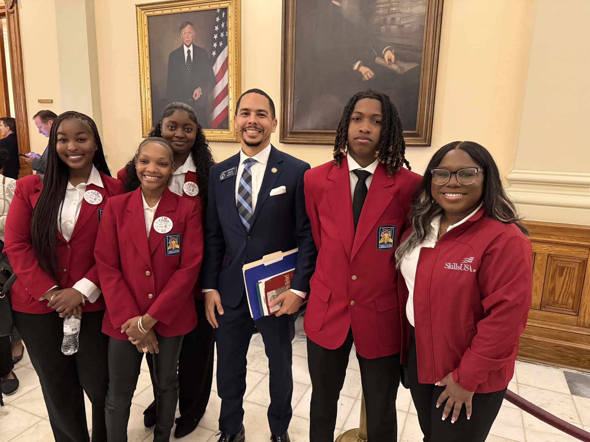 Five <span style='text-transform:none'><span style='text-transform:none'><span style='text-transform:none'>SkillsUSA</span></span></span> members in red blazers and an advisor in a red <span style='text-transform:none'><span style='text-transform:none'><span style='text-transform:none'>SkillsUSA</span></span></span> jacket pose with a state senator, whose name badge reads “Jaha Howard, Senator, District 39,” in a marble capitol hallway lined with framed portraits.” class=”wp-image-22321″/><figcaption class=