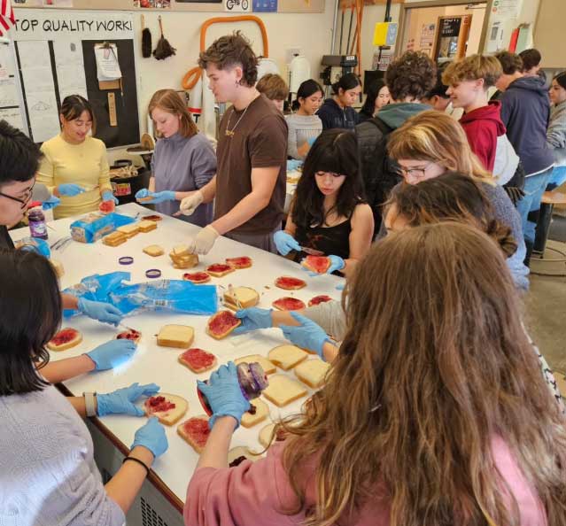 Students in blue gloves line both sides of a long table in a classroom, assembling peanut butter and jelly sandwiches on slices of bread, with loaves and jars spread across the table and more students gathered behind them.