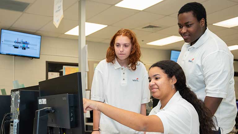 Three students in white SkillsUSA polo shirts gather at a computer workstation in a technical lab. One student is seated and points at the monitor while two others stand behind her and watch.